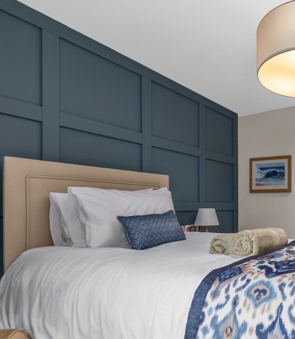 Bright, modern care home bedroom featuring a large bed with white linens, blue accent pillows and a patterned throw, a dark blue feature wall, soft overhead lighting, and an open door leading to an en-suite bathroom